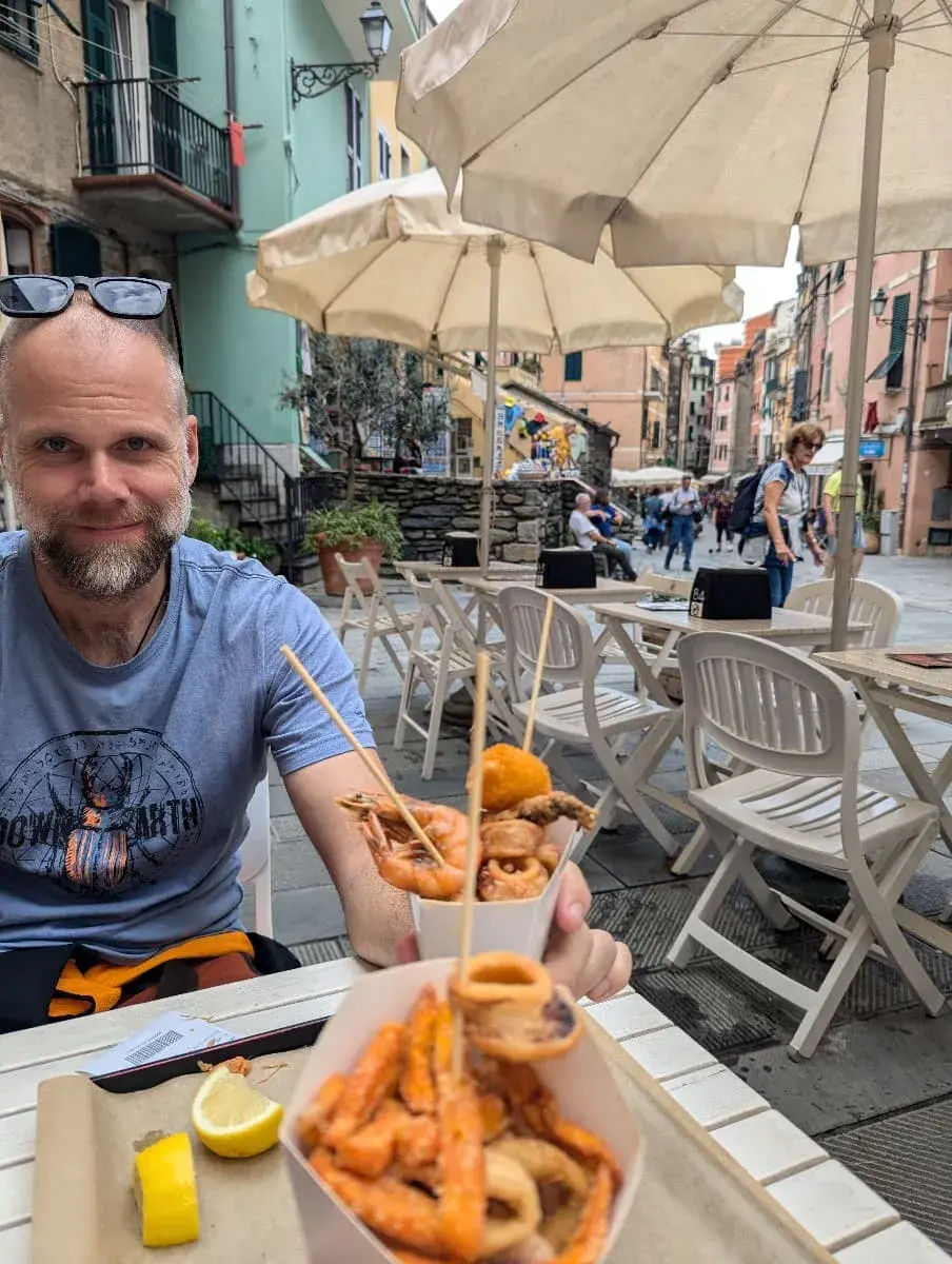 Jan sitzt in einem Straßencafé in Riomaggiore und hält eine Schale mit Fritto Misto, bestehend aus verschiedenen frittierten Meeresfrüchten. Im Hintergrund sind weitere Gäste und bunte Gebäude zu sehen.