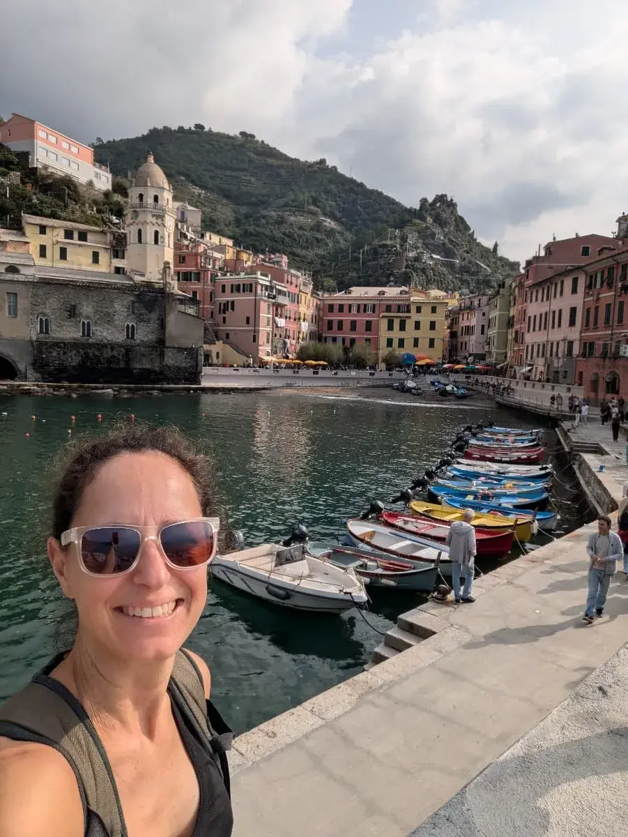 Ein Selfie von Linda mit Sonnenbrille im Hafen von Vernazza. Der kleine Hafen ist von bunten Häusern umgeben, und die charakteristische Kirche des Ortes mit ihrem Glockenturm ist prominent zu sehen.