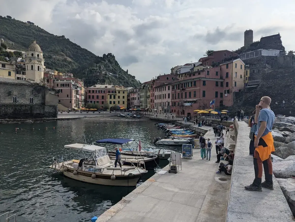 Der malerische Hafen in Vernazza, umgeben von bunten Gebäuden und einer Kirche mit Kuppel, mit vielen kleinen Booten im Wasser. Besucher schlendern entlang der Uferpromenade, während sich die Hügel der Cinque Terre im Hintergrund erstrecken.