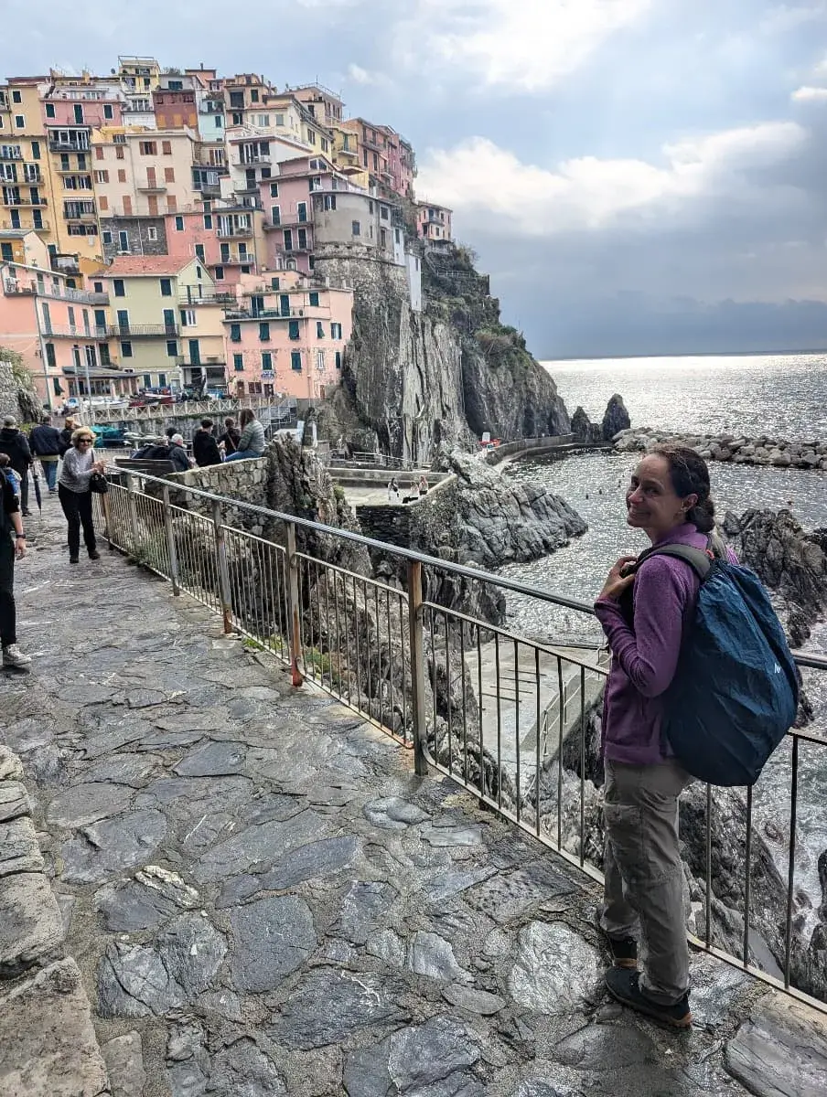 Linda mit Rucksack steht lächelnd am Rand eines steinigen Wegs, mit Blick auf die farbenfrohen Häuser, die sich an den steilen Klippen von Manarola aufreihen. Die Abendsonne lässt das Meer glitzern und taucht die Szenerie in warmes Licht.