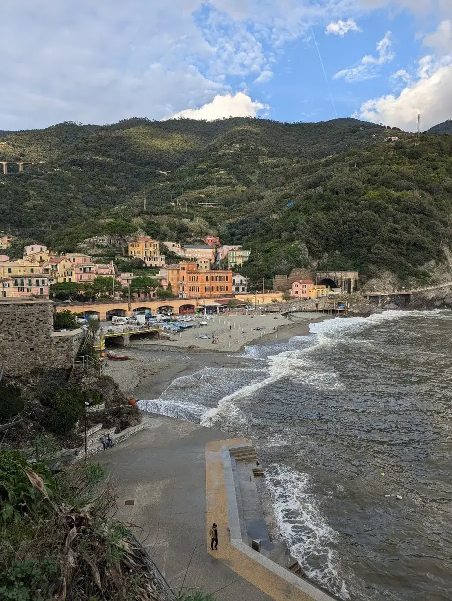 Die Küste von Monterosso al Mare ist gesäumt von pastellfarbenen Häusern und grünen Hügeln, während Wellen gegen den Strand schlagen. Ein Spazierweg entlang des Wassers führt zu bunten Sonnenschirmen und Strandliegen.