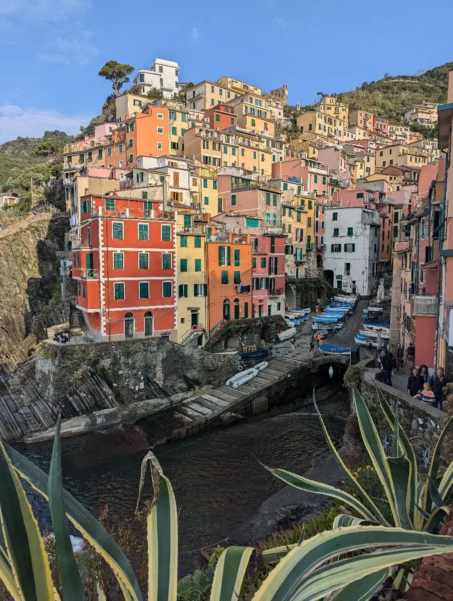 Die dicht aneinander gebauten, bunten Häuser von Riomaggiore im Cinque Terre Nationalpark spiegeln sich im ruhigen Wasser des kleinen Hafens. Kleine Fischerboote sind am Rand des Hafenbeckens vertäut, und eine steile Klippe bildet den Hintergrund.