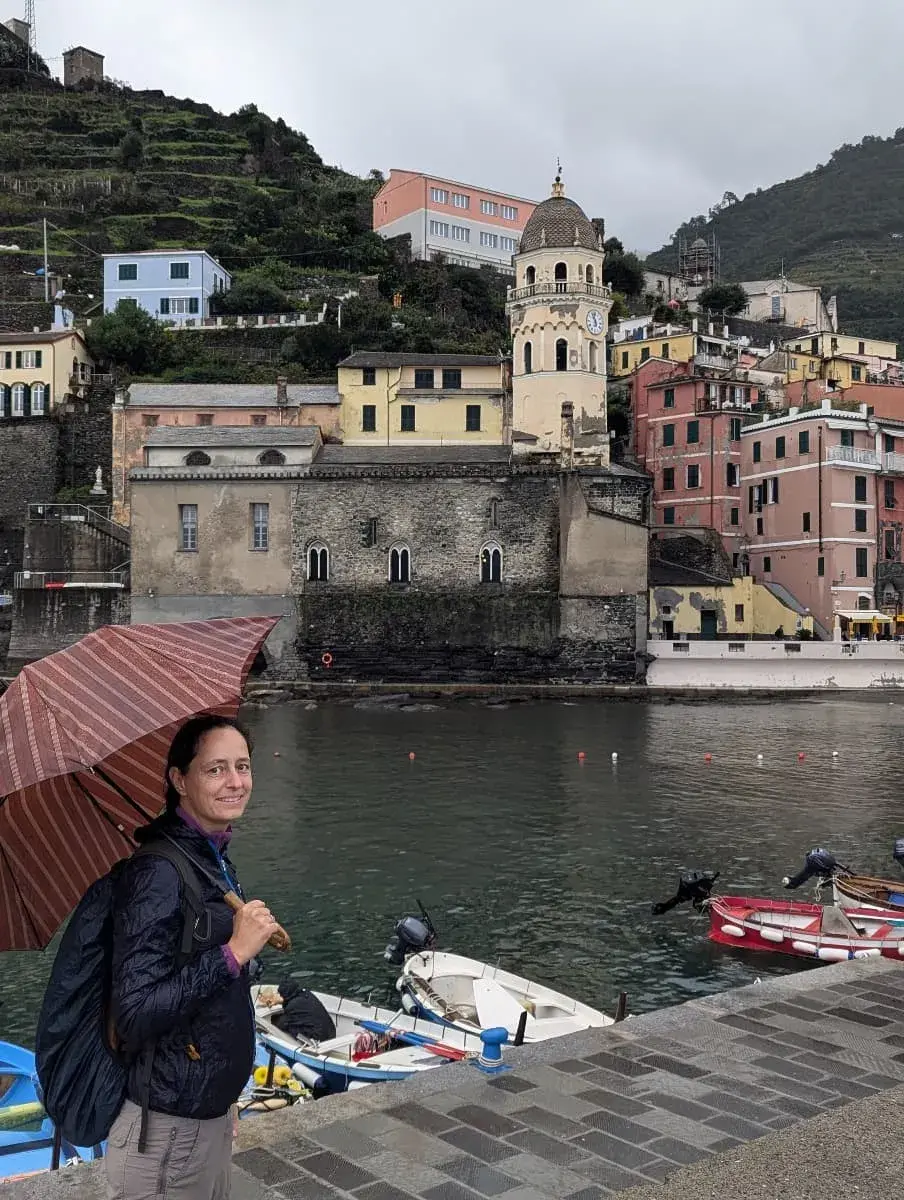 Linda mit Regenschirm spaziert entlang des Hafens von Vernazza, während ein Regenschauer das Küstendorf einhüllt. Die Kirche mit ihrem markanten Glockenturm und die farbenfrohen Häuser bilden einen stimmungsvollen Kontrast zur grauen Regenkulisse.
