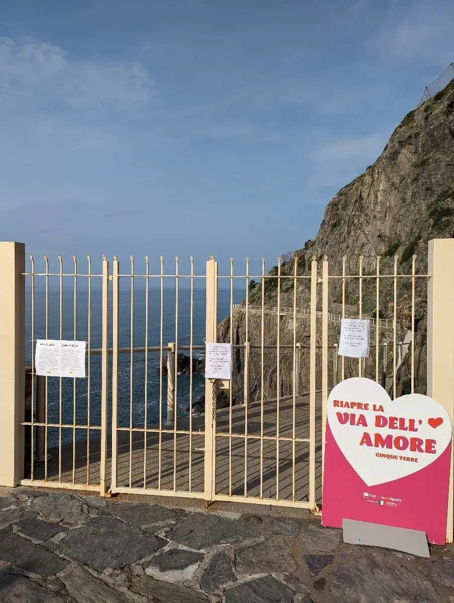 Ein verschlossenes Tor markiert den Eingang zur „Via dell’Amore“, einem berühmten Wanderweg entlang der Klippen. Ein großes Schild in Herzform weist auf die Wiedereröffnung des Weges hin, während sich der Weg im Hintergrund entlang der felsigen Küste schlängelt.