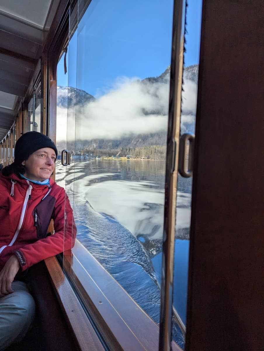 Linda schaut aus dem Fenster eines Ausflugsschiffs auf der Königssee Schifffahrt im Winter. Die Oberfläche des Königssees ist spiegelglatte, während sich Berge und Wolken im Hintergrund abzeichnen.