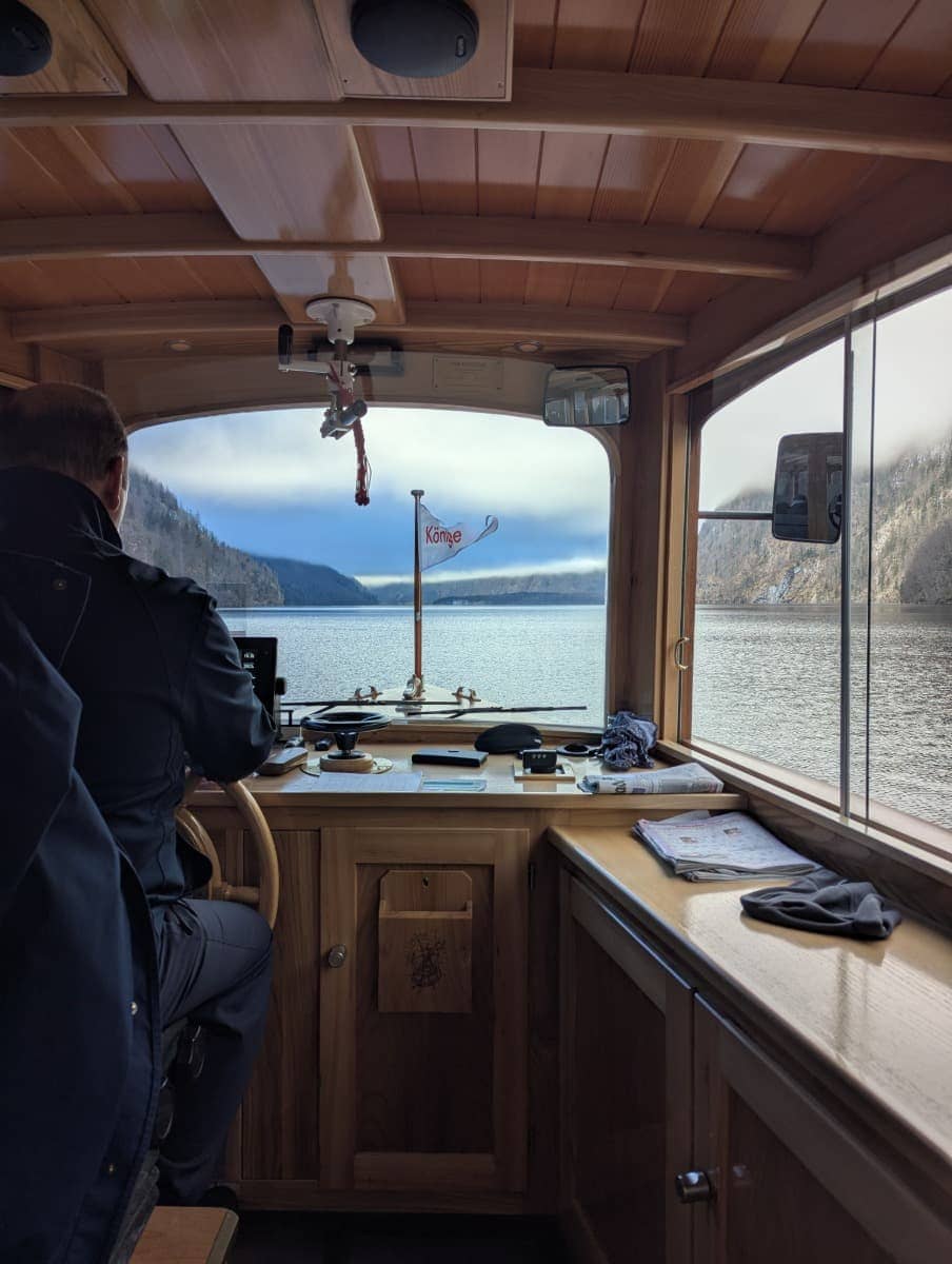 Innenansicht eines Ausflugsschiffs einer Königssee Schifffahrt im Winter, mit Blick auf den Kapitän am Steuerrad und den Ausblick auf das Wasser durch das Frontfenster.
