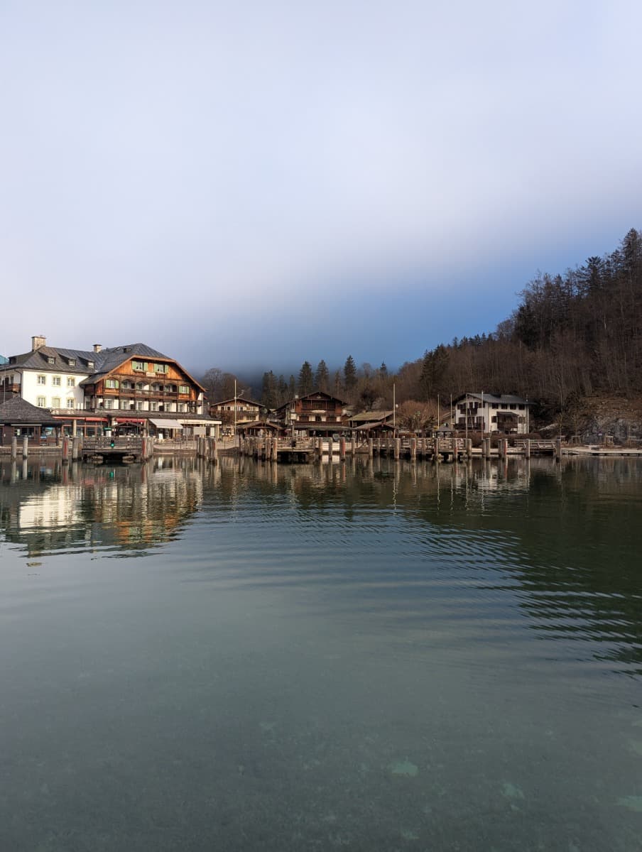 Die winterliche Anlegestelle in Schönau am Königssee mit traditionellen Gebäuden und stiller Wasseroberfläche, die das Dorf reflektiert.


