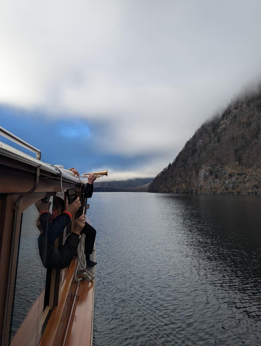 Der Kapitän spielt Trompete von der Seite eines Ausflugsschiffs auf dem Königssee, um das berühmte Echo des Sees zu erzeugen, eingerahmt von Bergen und Wolken.