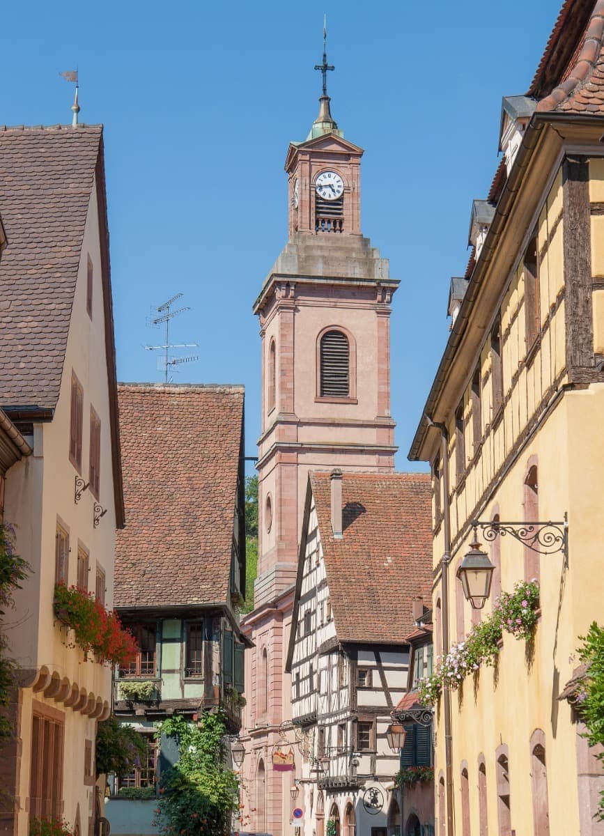 Das erste Bild zeigt eine malerische Gasse in Riquewihr, einem der schönsten Orte im Elsass, gesäumt von bunten Fachwerkhäusern mit blühenden Blumenkästen. Im Hintergrund ragt der Glockenturm der Kirche Saint-Jacques-le-Majeur in den strahlend blauen Himmel, während schmiedeeiserne Laternen die romantische Atmosphäre des Dorfes unterstreichen.