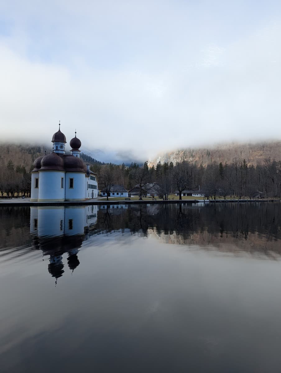 Die Kirche St. Bartholomä am winterlichen Königssee spiegelt sich im ruhigen Wasser, umgeben von dichtem Nebel und kahlen Bäumen. Die charakteristischen Zwiebeltürme stehen im Kontrast zur stillen, mystischen Winterlandschaft. 