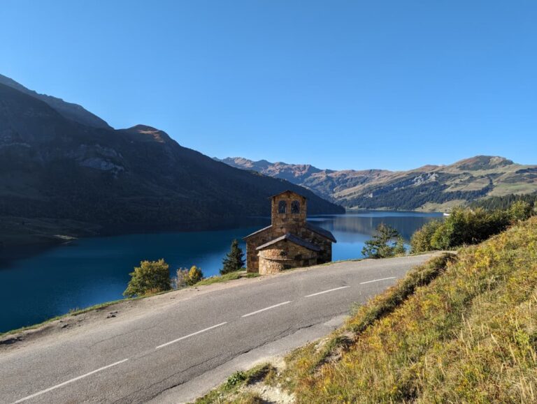 Eine kleine Steinkapelle steht idyllisch an einer geschwungenen Bergstraße mit Blick auf einen tiefblauen Bergsee. Die umliegenden sanften Berghänge und die ruhige Wasseroberfläche schaffen eine beeindruckende Kulisse. Diese Route gehört zu den schönsten Alpenpässen mit dem Auto und bietet atemberaubende Ausblicke auf die alpine Landschaft.