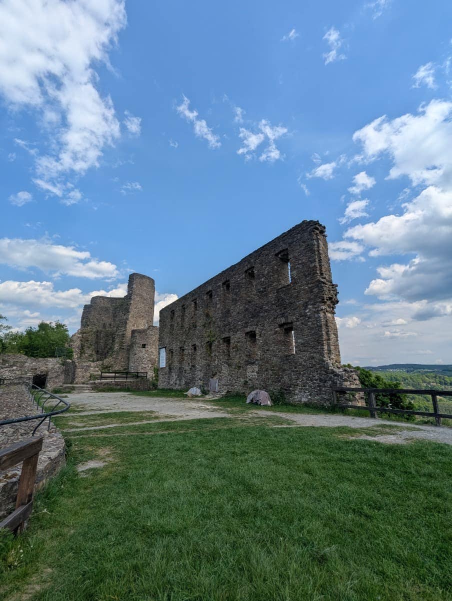 Die Ruine von Burg Windeck erhebt sich mit mächtigen Steinmauern unter einem wolkigen Himmel über grüner Wiese. Diese Kulisse zeigt eindrucksvoll, wie eindrucksvolle Kulturdenkmäler entlang des Natursteig Sieg ins Wandererlebnis eingebettet sind.