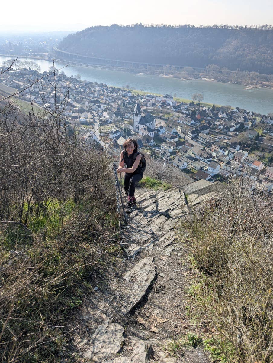 Linda erklimmt einen felsigen, steilen Pfad mit Wanderstöcken, im Hintergrund liegt Leutesdorf am Rhein. Der steinige Abschnitt macht die sportliche Herausforderung mancher Rheinsteig-Passagen spürbar.