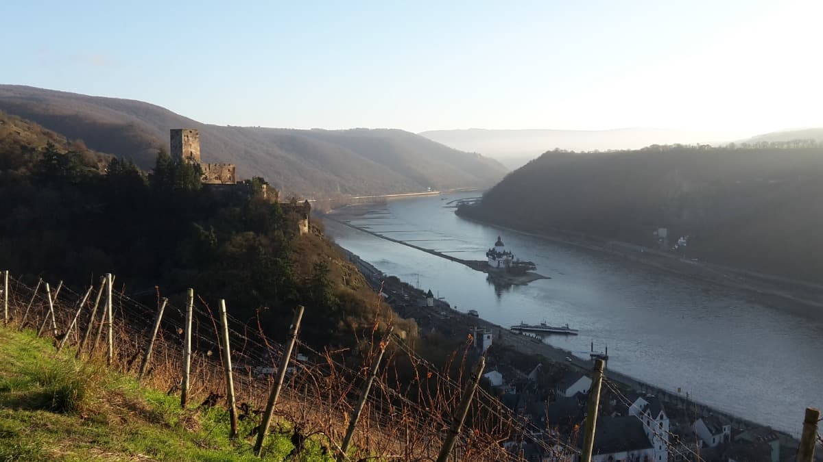 Die Szene zeigt eine Burg auf einem Hügel, die Burg Pfalzgrafenstein mitten im Rhein sowie das Dorf Kaub in warmem Abendlicht. Das Foto unterstreicht die kulturellen Höhepunkte entlang des Rheinsteigs und passt zu Hinweisen auf geschichtsträchtige Etappen.