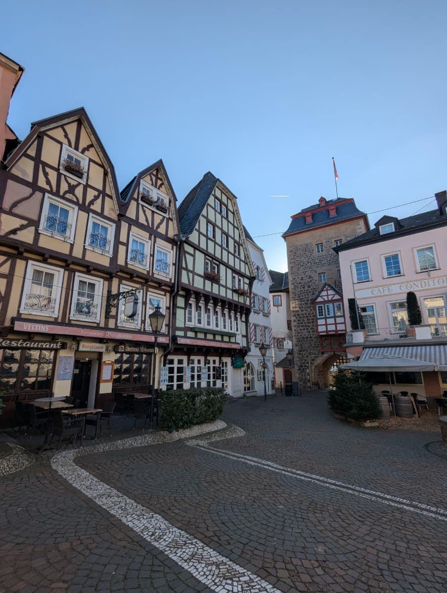 Mehrere bunte Fachwerkhäuser säumen einen ruhigen, kopfsteingepflasterten Platz mit Cafés und Gasthäusern. Die Szene spiegelt das historische Flair kleiner Rheinorte wider, durch die der Wanderweg führt.