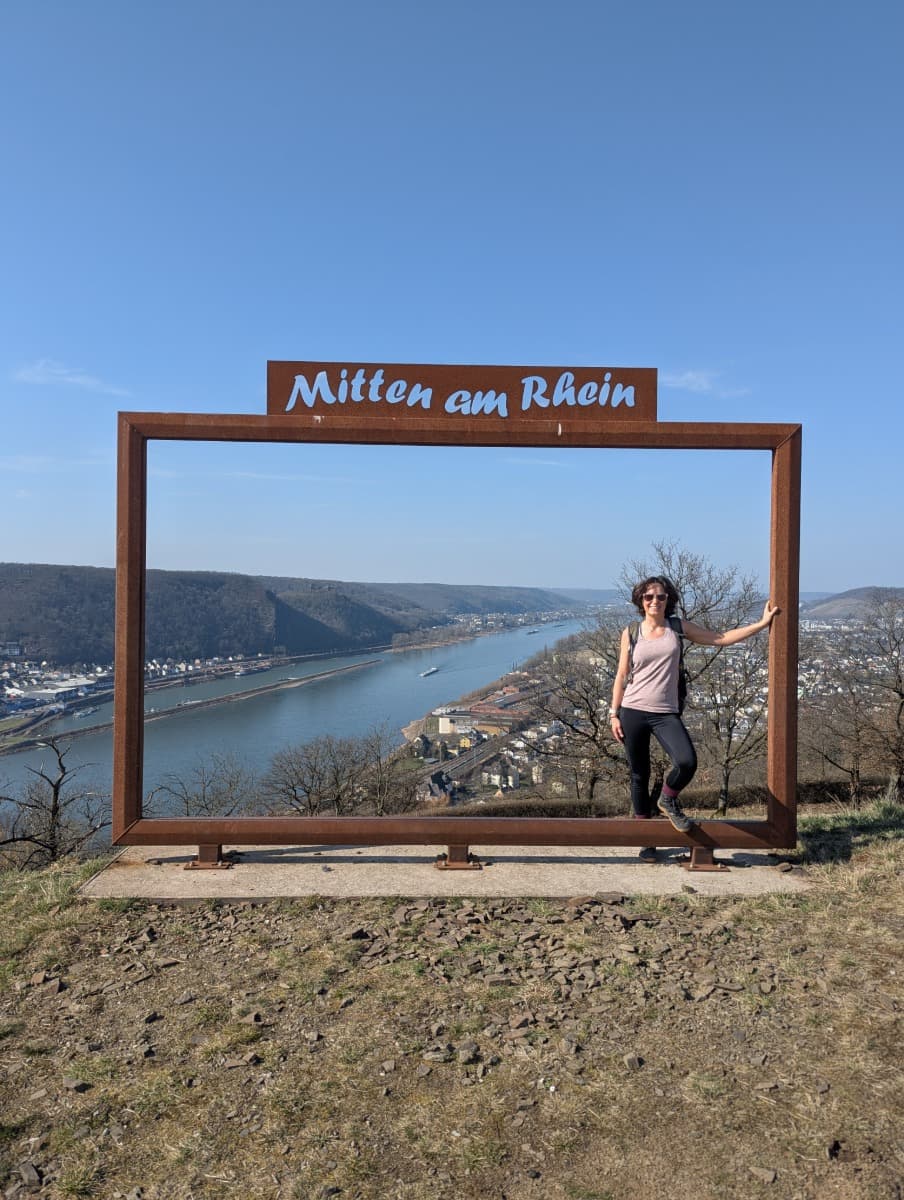 Linda steht in einem großen Bilderrahmen mit der Aufschrift „Mitten am Rhein“, dahinter fließt der Rhein durch ein weites Tal. Dieses Bild hebt einen markanten Fotospots am Rheinsteig hervor, der sich für unvergessliche Erinnerungsfotos eignet.