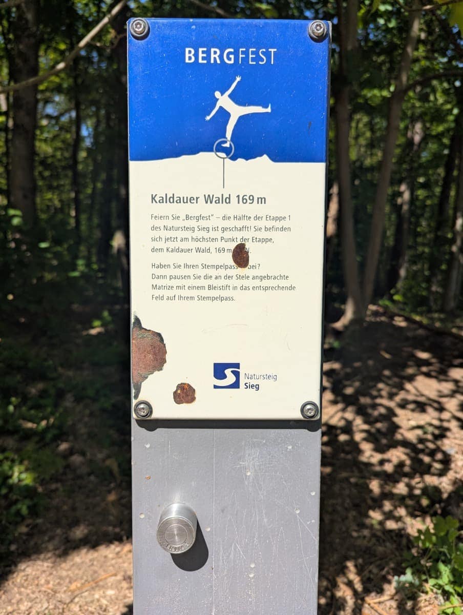 Ein blauer Wegweiser mit der Aufschrift „Bergfest“ markiert den höchsten Punkt der Etappe 1 im Kaldauer Wald bei 169 m Höhe. Die Szene verdeutlicht, wie der Natursteig Sieg das Wandern mit spielerischen Stempelstationen auflockert.