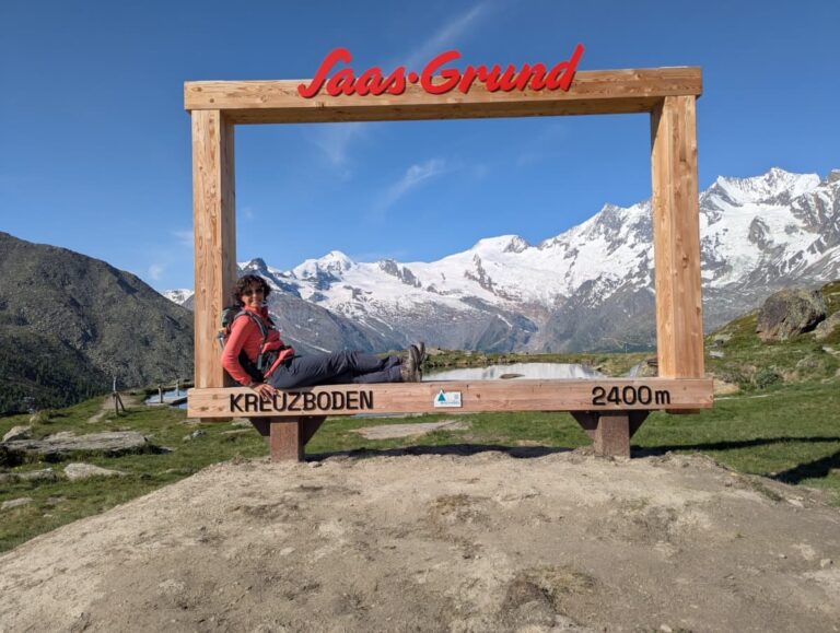Linda liegt entspannt im großen Holzrahmen am Aussichtspunkt Kreuzboden auf 2400 Metern – mit Panorama über das Saastal und seine majestätischen Gletscher, ideal zum Erinnerungsfoto beim Wandern im Saastal.