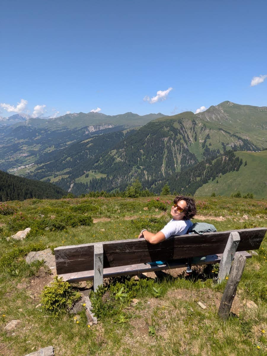 Linda auf einer Holzbank mit Blick auf die umliegenden Berge und Täler unter blauem Himmel. Erholsamer Moment beim Wandern in Arosa.