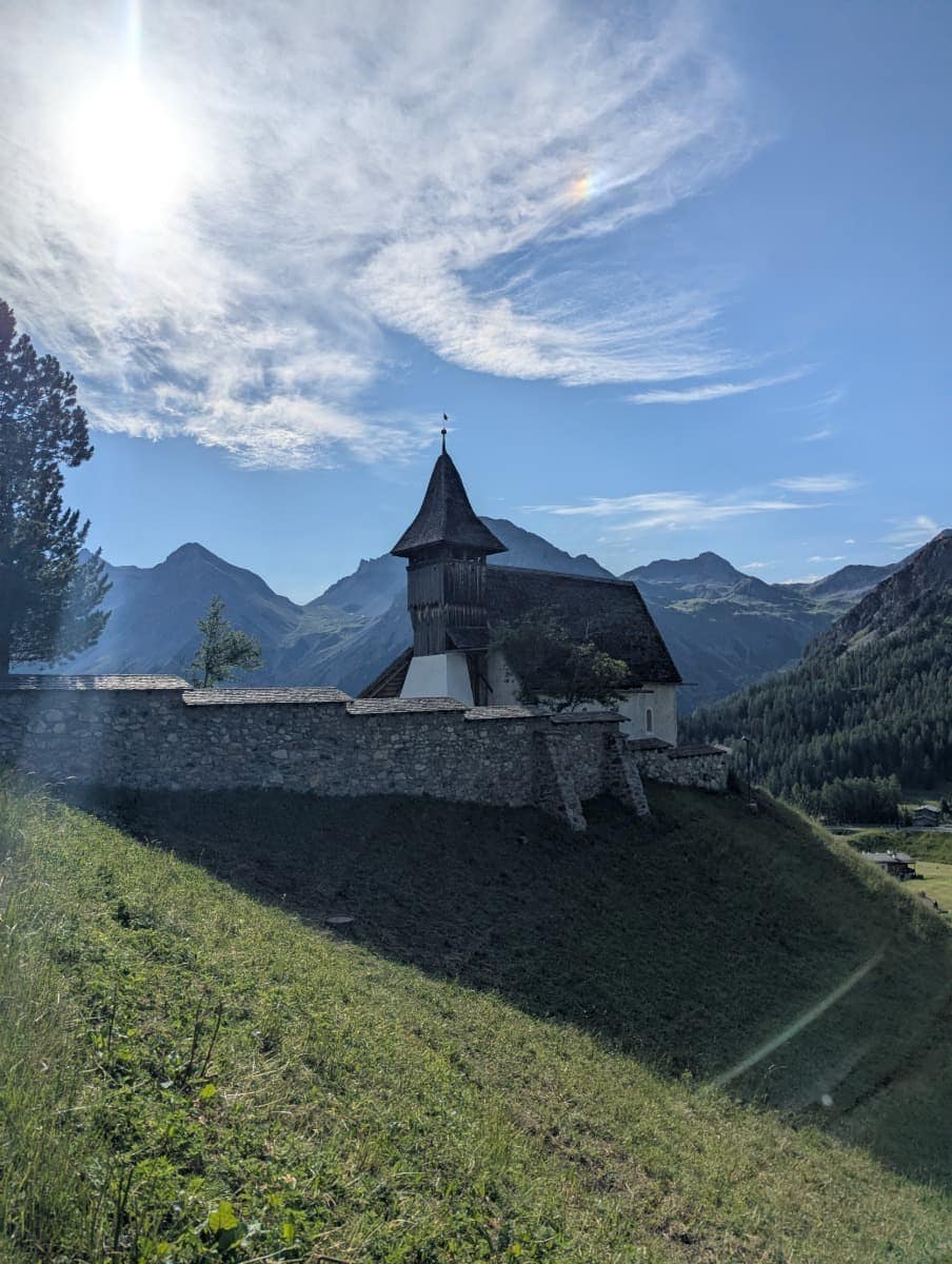 Alte Kirche mit Schindeldach auf einem Hügel mit Blick auf das Schanfigg Tal von Arosa, dahinter die Alpen im Gegenlicht. Historischer Zwischenstopp beim Wandern in Arosa.