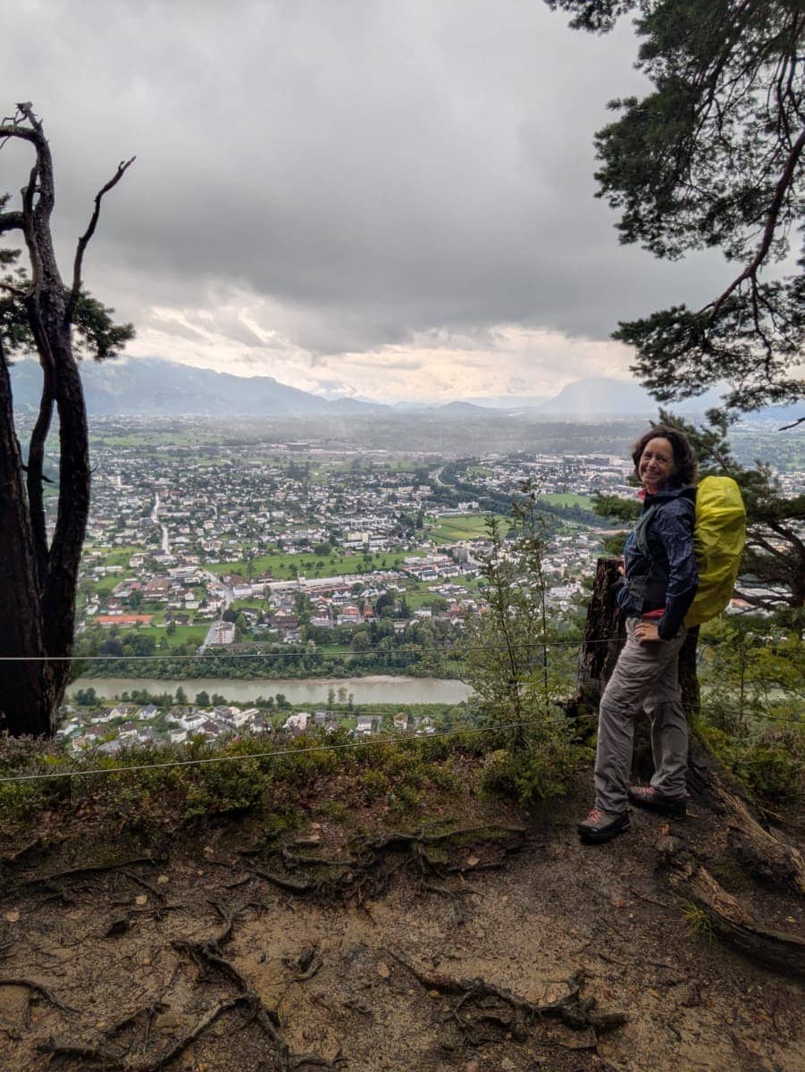 Linda steht mit gelber Regenhülle auf dem Rücken am Aussichtspunkt Känzele, mit weitem Blick über Bregenz, den Rhein und den Bregenzerwald unter grauem Himmel. Auch bei Regen lohnt sich dieses Ausflugsziel von Dornbirn in Vorarlberg.