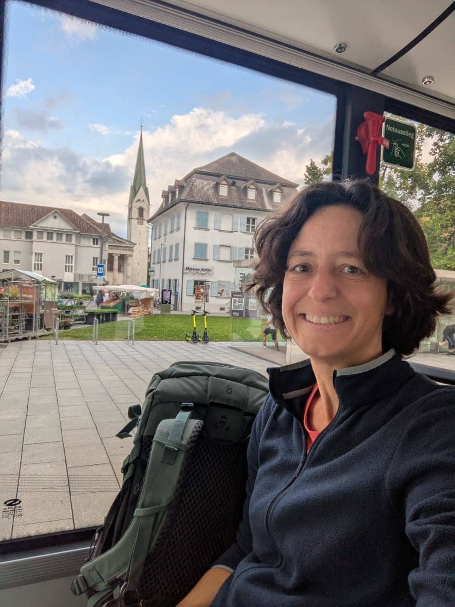 Linda sitzt lächelnd mit Wanderrucksack im Bus, im Fenster spiegeln sich ein Kirchturm und Altbauten der Dornbirner Innenstadt. Öffentliche Verkehrsmittel sind beim Dornbirn Kurztrip praktisch und bequem.
