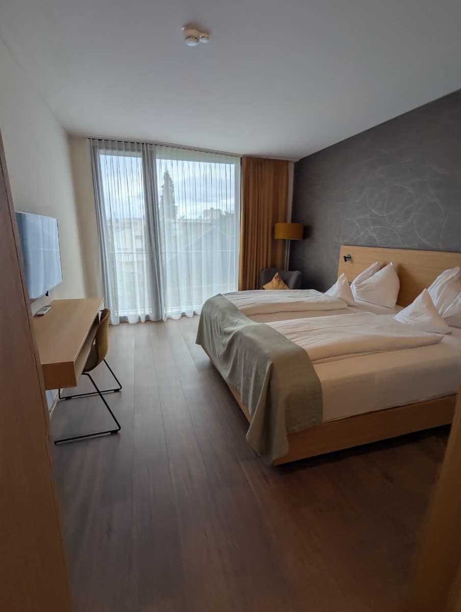 Helles Hotelzimmer mit Doppelbett, Schreibtisch, TV und raumhohem Fenster mit halbtransparentem Vorhang. Nach einem aktiven Tag in Dornbirn in Vorarlberg lässt es sich hier gemütlich entspannen.