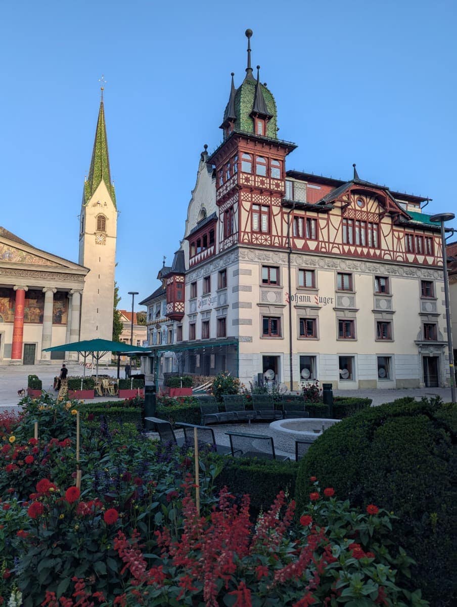 Blühende Beete und Sitzbänke vor dem prächtigen Gebäude der Sparkasse Dornbirn mit Fachwerk und Türmchen, daneben die Stadtpfarrkirche St. Martin. Der Marktplatz von Dornbirn in Vorarlberg ist ein echtes Postkartenmotiv.