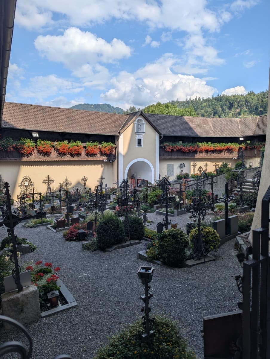 Blick auf den gepflegten Klosterfriedhof der Basilika Rankweil, mit schmiedeeisernen Grabkreuzen, bunten Blumen und einem Torbogen, eingerahmt von Bergen. In der Umgebung von Dornbirn in Vorarlberg warten ruhige, kulturreiche Orte wie dieser.