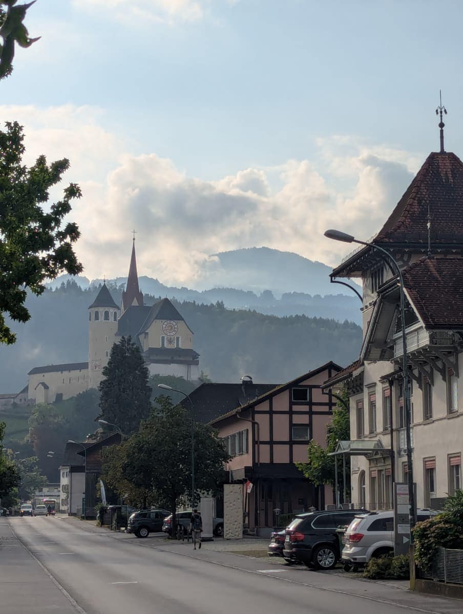 Die Rankweil Basilika mit roten Türmen thront über der kleinen Stadt in Vorarlberg, umgeben von Nebel, alten Häusern und sanften Hügeln im Hintergrund. Auf einem Dornbirn Kurztrip lassen sich auch umliegende Orte mit historischen Zentren entdecken.