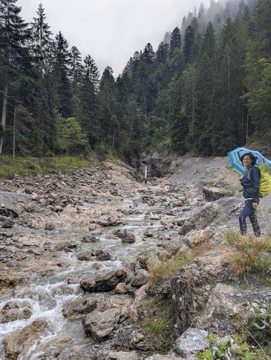 Linda steht mit Regenschirm unterhalb von Buchboden an einem Wildbach, der sich durch ein steiniges Flussbett schlängelt, umgeben von dichtem Wald und Nebelschwaden. Auch bei grauem Wetter ist das Große Walsertal ein beeindruckendes Naturerlebnis.