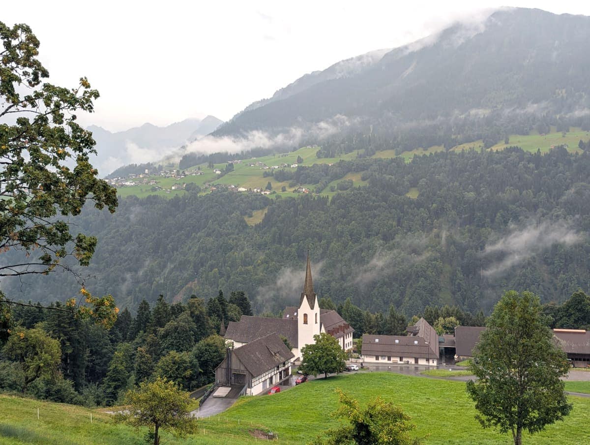 Ein Blick auf die Propstei St. Gerold mit Kirche und Nebengebäuden, eingerahmt von nebelverhangenen Hängen und verstreuten Häusern auf der gegenüberliegenden Talseite. Diese Szenerie zeigt die typische Streusiedlung im Großen Walsertal.