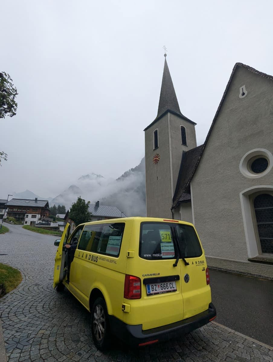 Ein gelber Landbus mit der Aufschrift „Großes Walsertal“ steht vor der Kirche in Buchboden mit spitzem Turm und Sonnenuhr, während sich Nebel über die Dächer im Hintergrund zieht. Auch ohne Auto ist das Große Walsertal gut mit öffentlichen Verkehrsmitteln zu erkunden.
