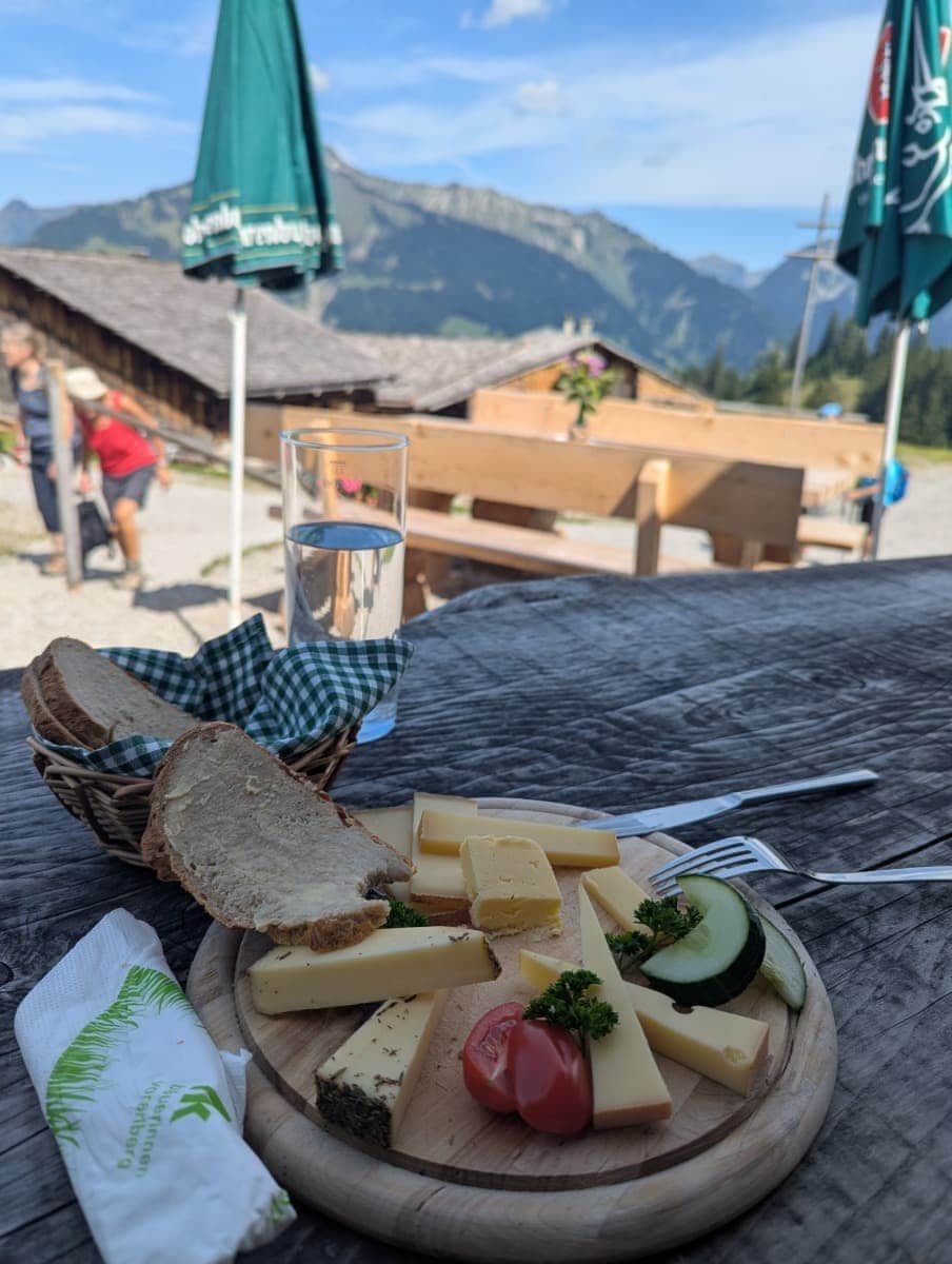 Auf einem Holzteller liegen verschiedene regionale Käsesorten, Brot, Tomaten und Gurken, dahinter eine Holzhütte mit Sonnenschirmen und Bergblick. Kulinarischer Genuss gehört im Großen Walsertal genauso dazu wie die Natur.