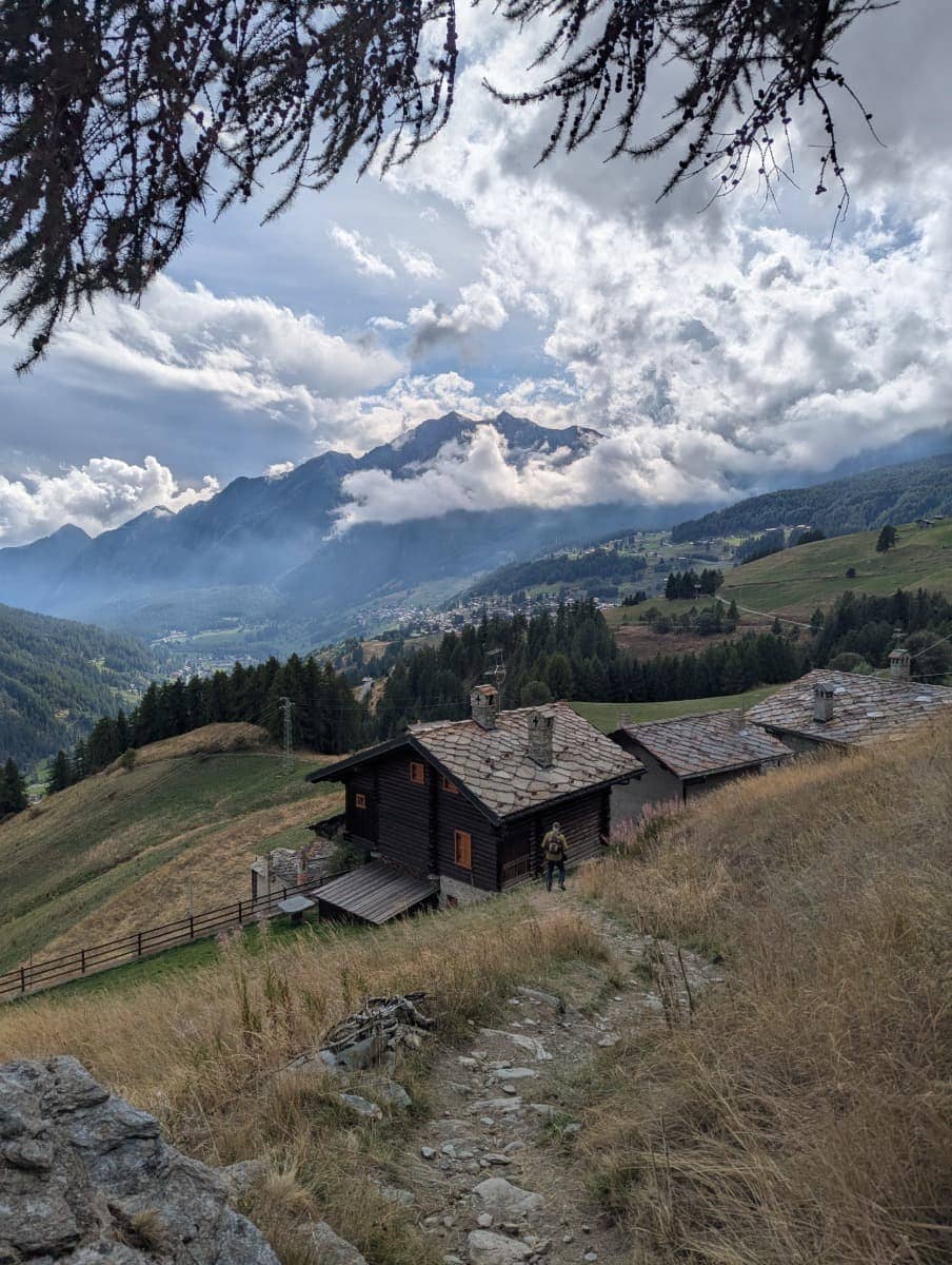 Ein traditionelles Steinhaus im Ayastal liegt idyllisch zwischen Almwiesen mit Blick ins Aostatal. Die Szenerie zeigt die typische Architektur und die weite Berglandschaft.