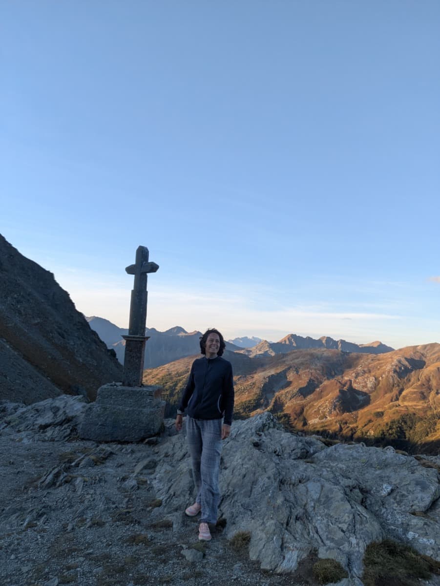 Linda steht am Gipfelkreuz auf dem Großen Sankt Bernhard Pass und blickt über die Bergketten des Aostatals, die in goldenes Morgenlicht getaucht sind.