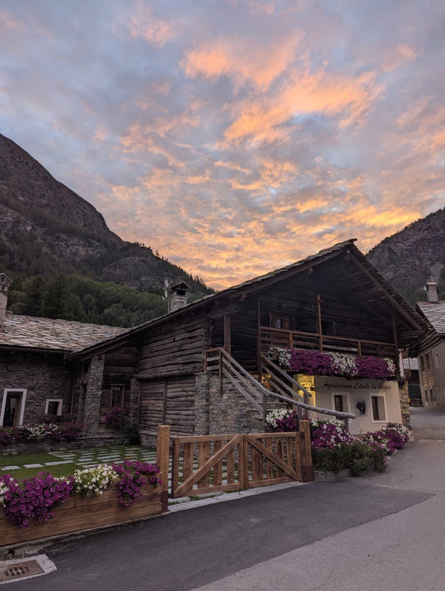 Ein traditionelles Steinhaus in Cogne, im Weiler Lillaz, ist mit Blumen geschmückt, während über den Bergen ein farbenprächtiger Abendhimmel leuchtet. Das Bild des Agriturismo L'Etoile du Berger unterstreicht die alpine Stimmung und den Charme der Unterkünfte im Aostatal.