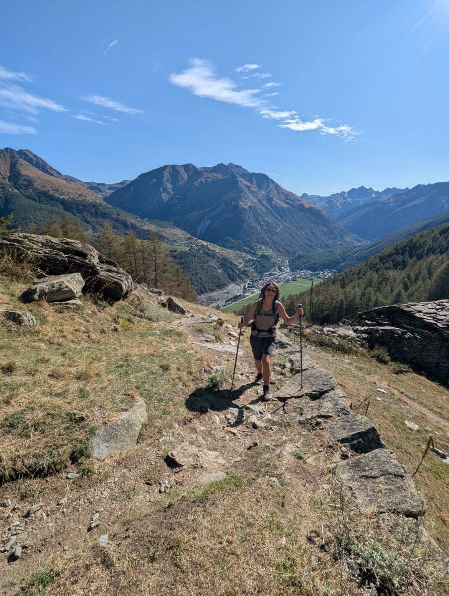 Linda wandert mit Stöcken einen steinigen Pfad oberhalb von Cogne zum Weiler Les Ors-Dessus hinauf. Hinter ihr liegen das Cogne-Tal und Berge. 
