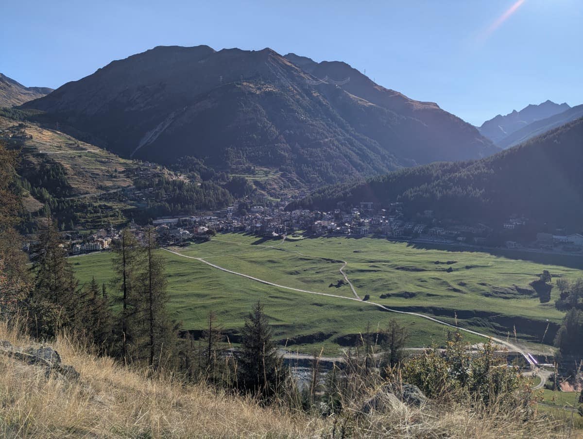 Über der weiten Wiesenlandschaft von Cogne erhebt sich eine markante Bergkette, im Vordergrund führen Wanderwege durchs Grün. Diese Ansicht zeigt die offene Tallage und das Bergpanorama des Ortes.