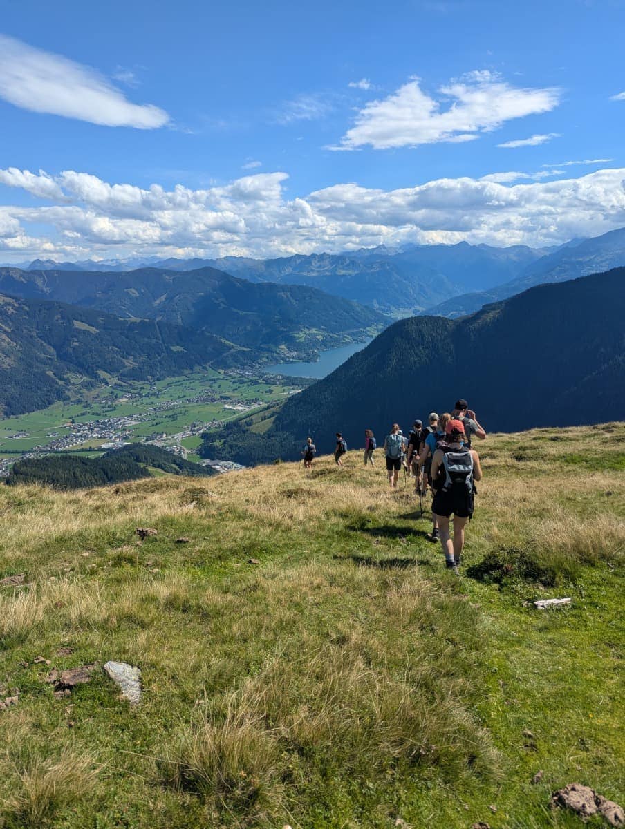 Eine Gruppe Wanderer steigt über eine Wiese hinab, im Hintergrund glitzert der Zeller See zwischen den Bergen. Die Szene zeigt die Vielfalt der Ausblicke entlang des Saalachtaler Höhenwegs.