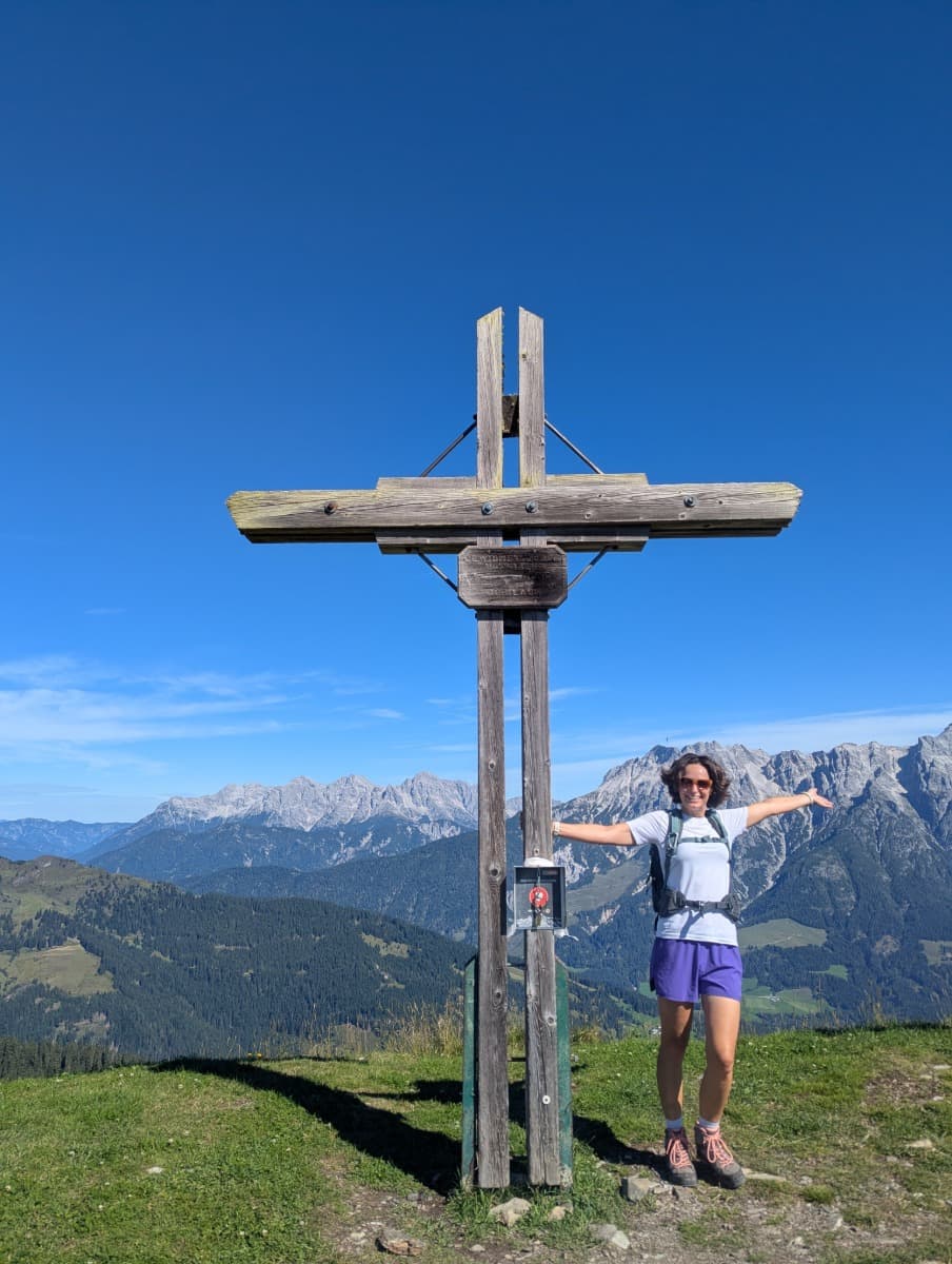 Linda steht mit ausgebreiteten Armen am Gipfelkreuz des Wildenkarkogel, hinter ihr das Steinerne Meer. Das Bild unterstreicht die Freude am Erreichen der Höhenziele auf dem Saalachtaler Höhenweg.