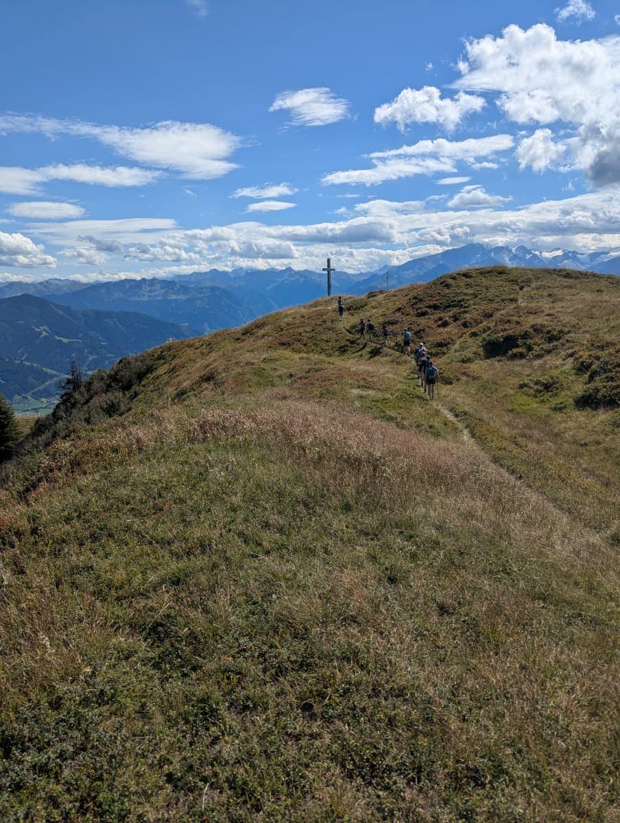 Wanderer nähern sich über einen grasbewachsenen Rücken dem Sausteige Gipfelkreuz. Die Wolken ziehen über die Bergspitzen. Hier wird die Weitläufigkeit und Ursprünglichkeit des Saalachtaler Höhenwegs sichtbar.
