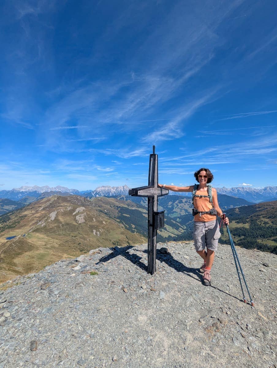 Linda steht mit ihren Wanderstöcken stolz am Gipfelkreuz des Saalbachkogels. Von hier eröffnet sich ein 360-Grad-Panorama über das Glemmtal und die Kitzbüheler Alpen.