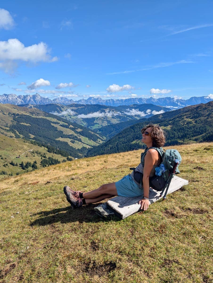 Linda macht eine Pause auf dem Pinzgauer Spaziergang oberhalb von Saalbach Hinterglemm. Das Panorama reicht über grüne Täler bis zu den Gipfeln der Kitzbüheler Alpen.