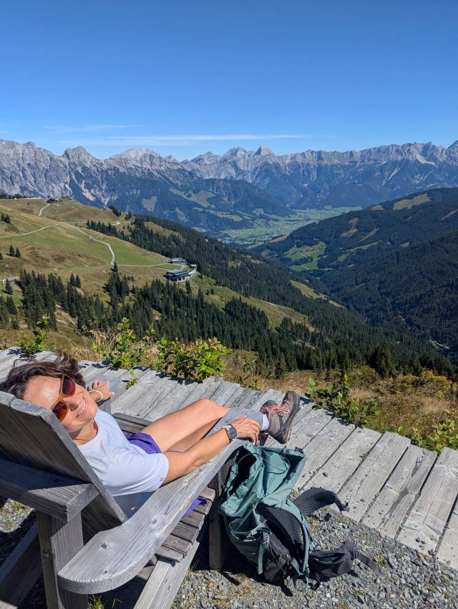 Linda entspannt auf einer Holzliege mit Blick ins Tal und die Leoganger Steinberge am Saalachtaler Höhenweg. Das Alpenkino ist nur eines der Highlights auf einem der schönsten Höhenrouten bei Saalbach Hinterglemm.