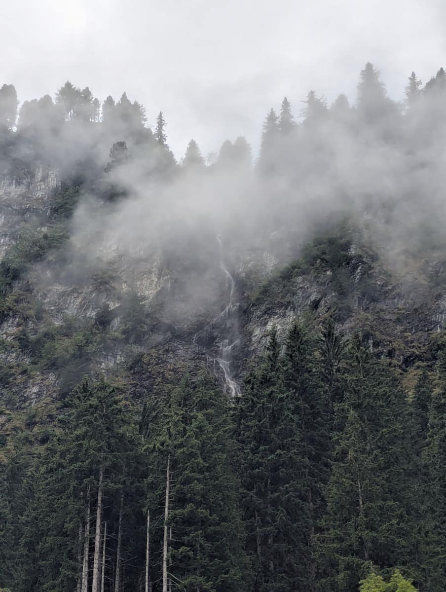 Ein Wasserfall stürzt durch Nebelschwaden über eine Felswand oberhalb von Hinterglemm. Das Naturmotiv zeigt die wilde Seite der Berge auf dem Talschluss Höhenweg.