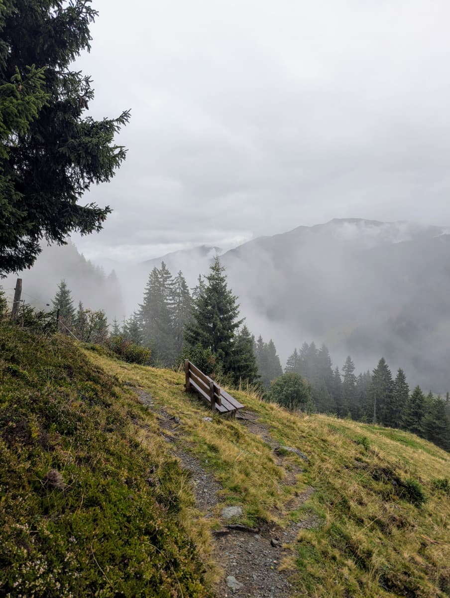 Eine Bank steht am Waldrand im Nebel mit Ausblick ins Glemmtal. Sie liegt am Talschluss Höhenweg, wo auch an trüben Tagen die Bergstimmung spürbar wird.