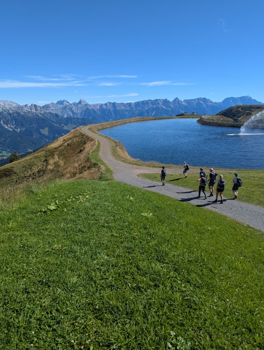 Wanderer spazieren am Speichersee entlang des Saalachtaler Höhenwegs mit Sicht auf die Leoganger Steinberge. Der künstliche See ist ein markanter Orientierungspunkt am Panoramaweg.