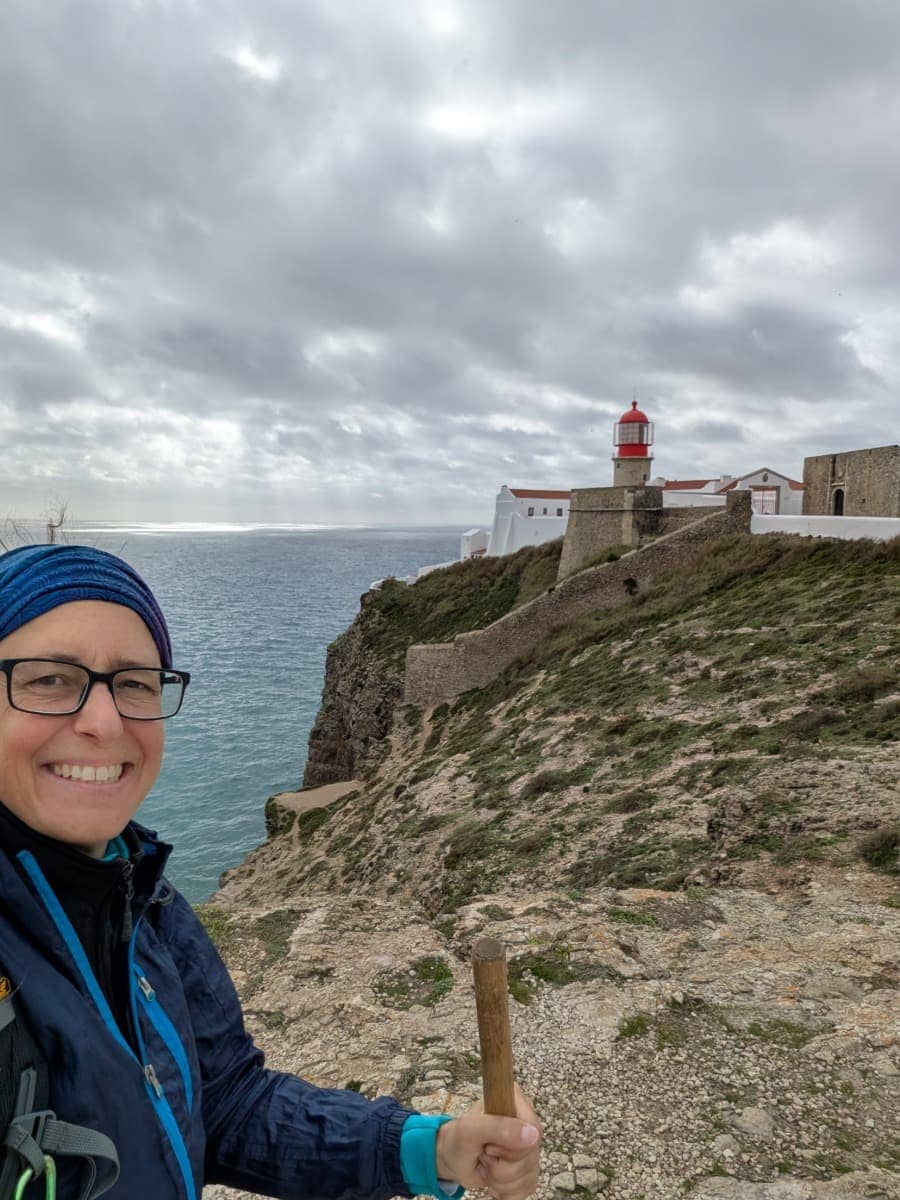 Linda steht mit Wanderstock auf der felsigen Klippe am Cabo de São Vicente, während hinter ihr der rot-weiße Leuchtturm und die weite Atlantikküste unter dichtem Wolkenhimmel liegen. Der markante Aussichtspunkt an der Südwestspitze Portugals zeigt, wie eindrucksvoll Weitwandern im Winter entlang des Fischerweges mit rauer Küste und weitem Meerblick sein kann.