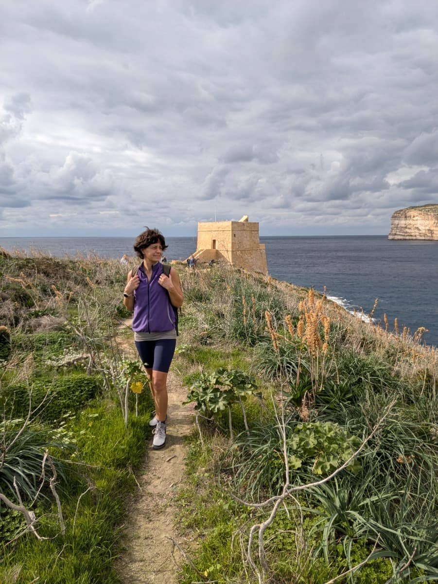 Linda wandert mit Rucksack auf einem schmalen Gozo Coastal Walk durch grüne Vegetation, im Hintergrund steht ein historischer Wachturm direkt über dem Meer. Das Motiv zeigt Weitwandern im Winter auf Malta, wo gut begehbare Küstenwege und milde Temperaturen auch außerhalb der Saison ideale Bedingungen schaffen.